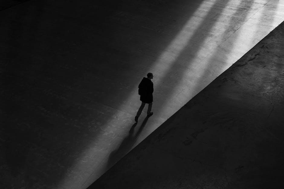 Black and white photo of a man walking through shadows on a deserted pavement, creating a moody silhouette.